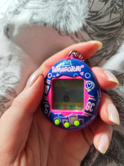 photograph of a white hand holding a 'tamagotchi'- and egg shaped pixel game with a small character.