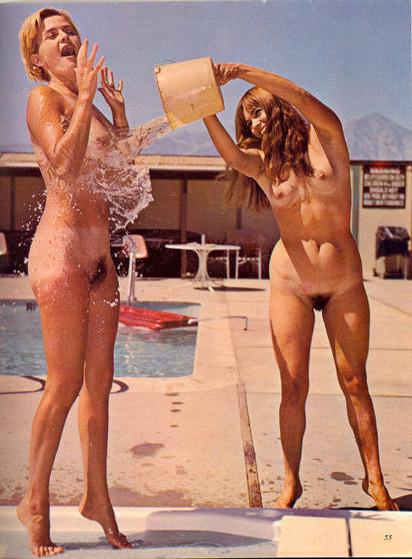 An old color photo of a nude woman throwing a bucket of water onto another nude woman next to an outdoor swimming pool.