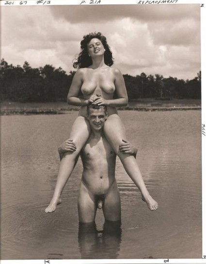 An old black and white photo of a nude woman riding atop a nude man's shoulders. The man stands in calm, thigh-deep water.