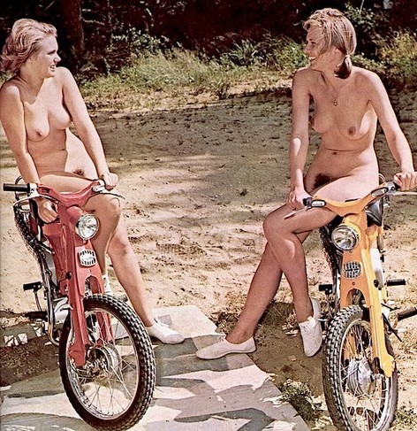 An old color photo of two nude women sitting atop mopeds facing one another.