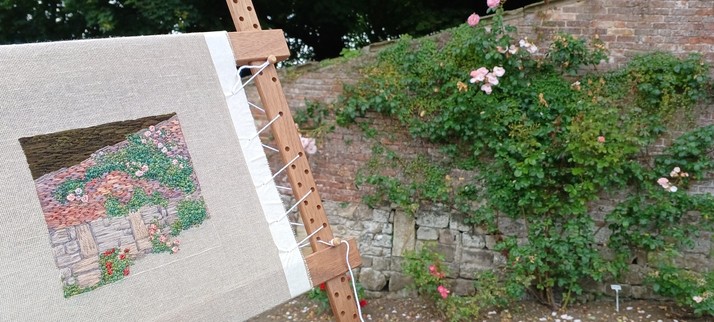 A stone and brick wall with a climbing roses. A part worked stitched artwork depicting the wall is held up in front and to the left