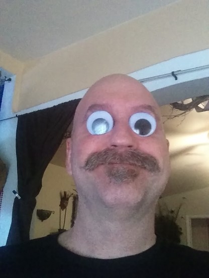 A color photograph of a man with a shaved head, mustache and soul patch who has two large googly eyes being held in his eye sockets. Oh and he has a goofy smile.