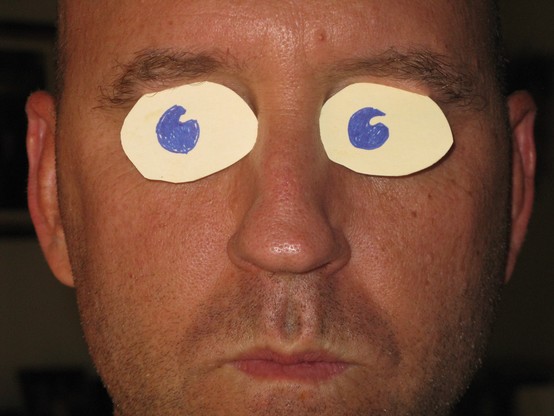A color photograph, selfie, of a man with two hand drawn eyes cut from card stock and placed in his eye sockets. He is otherwise serious looking.