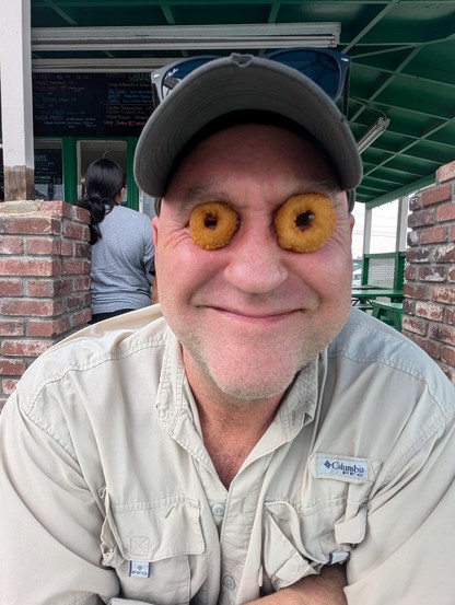 A color photograph of a man in a hat who has taken two different sized onion rings and placed them over his eyes. He is back to that same goofy smile and if he had realized he would be picking pieces of onion ring our of his eyes for the next hour wouldn't have done this.