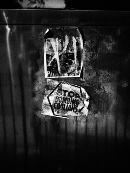 Weathered stickers on the side of an electrical box illuminated by diffuse light through a grated metal fence. One of the stickers says “stop bombing civilians”