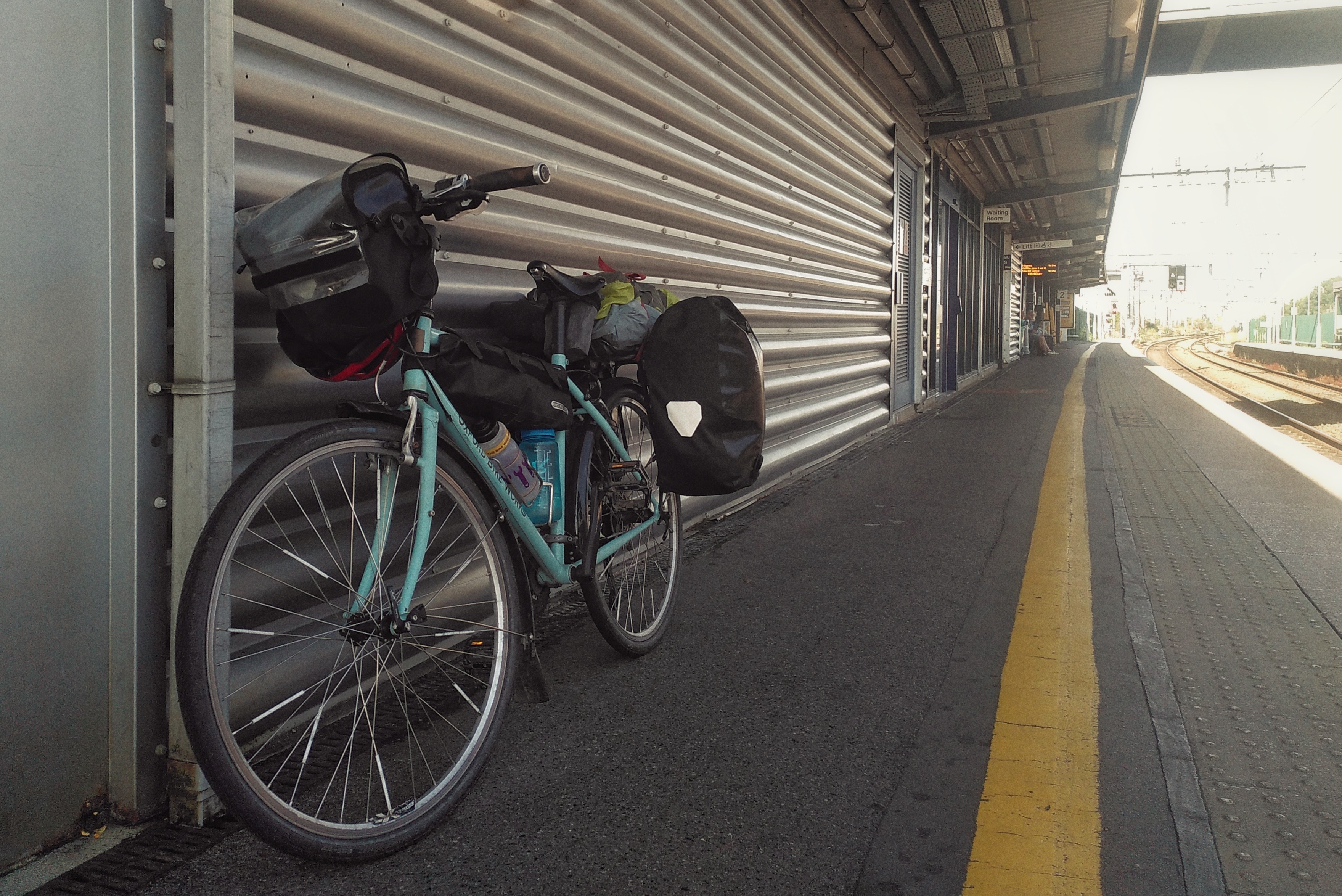 A horizontal colour photograph of a bicycle resting on a metal wall by an empty train platform.