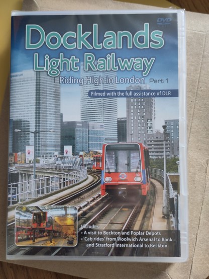 DVD called Docklands Light Railway: Riding High in London Part 1. Cover shows a red DLR train on a raised concrete railway, behind are the skyscrapers of the Canary Wharf development. An inset shows a jacked-up train in a depot. Straplines promise can rides and a depot visit, all 'filmed with the full assistance of DLR'.