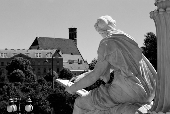 The black and white image shows a statue of a seated woman viewed from behind. She holds a writing board in her left hand and is dressed in flowing garments. The statue is positioned outdoors, with a background of trees and a large, old building with a steep roof and a tall tower. The sky is clear and sunny.