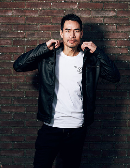 Man in a leather jacket and white t-shirt against a brick wall.