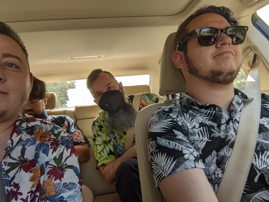 Four white people with brown or brown and blue hair and dark facial hair in tropical patterned short sleeved button downs in a car. 