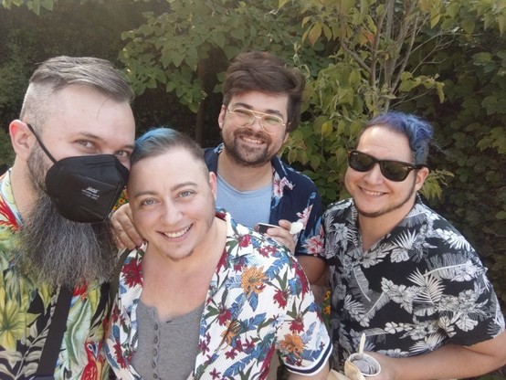Four white people with brown or brown and blue hair and dark facial hair in tropical patterned short sleeved button downs in a garden. 