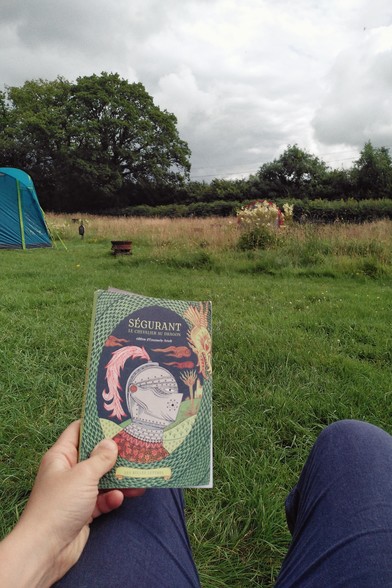 A vertical colour photograph of my hand above my knees holding up a book (Ségurant le chevalier au dragon edired by Emanuele Arioli) in a lush campsite. The sky is covered on grey clouds.