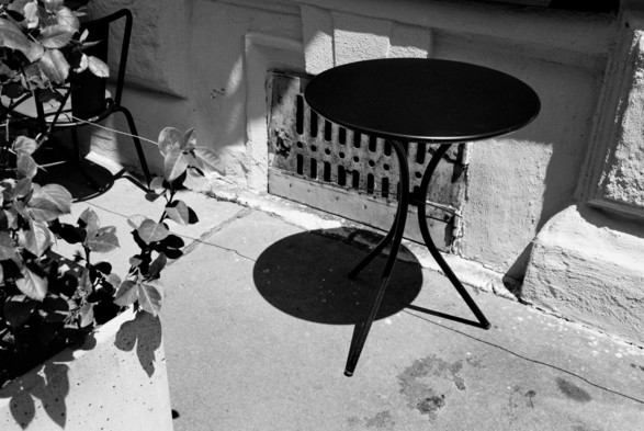 The black and white image shows a small round table with three legs placed on a sidewalk next to a building. There is a potted plant with leafy branches on the left side of the table. The table casts a shadow on the ground, and the building wall behind it has a ventilation grate.
