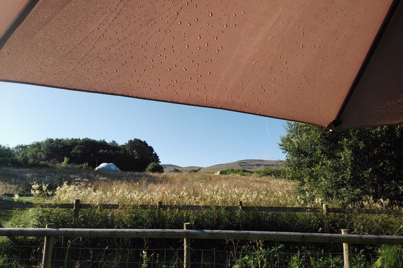 A horizontal colour photograph of a camping field under blue sky. The top of the image is obstructed by part of a sun umbrella.