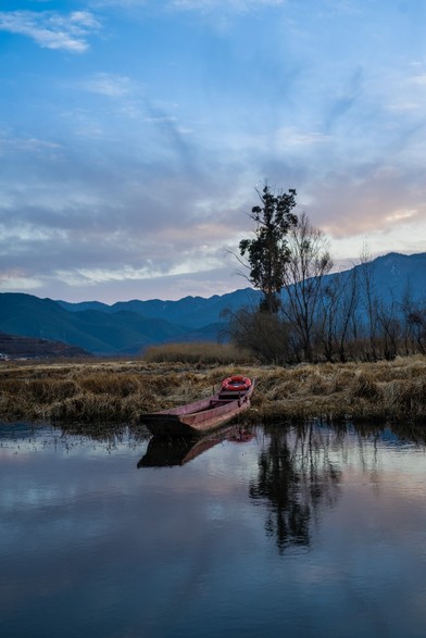 Color photo centered around a wooden rowing boat at the shore of a lake. It's just after sunset and the warmer tones of the wood are offset against the blue haze that encompasses the mountain range at the horizon.