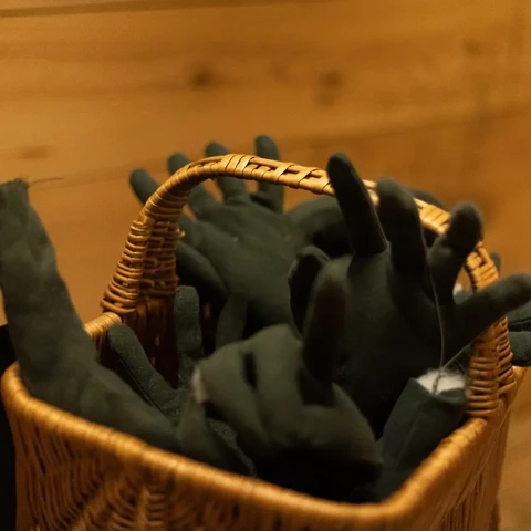 A handbasket with a bunch of green severed hands (fabric) in front of a wood panel wall. The Handbasket is part of an art installation.