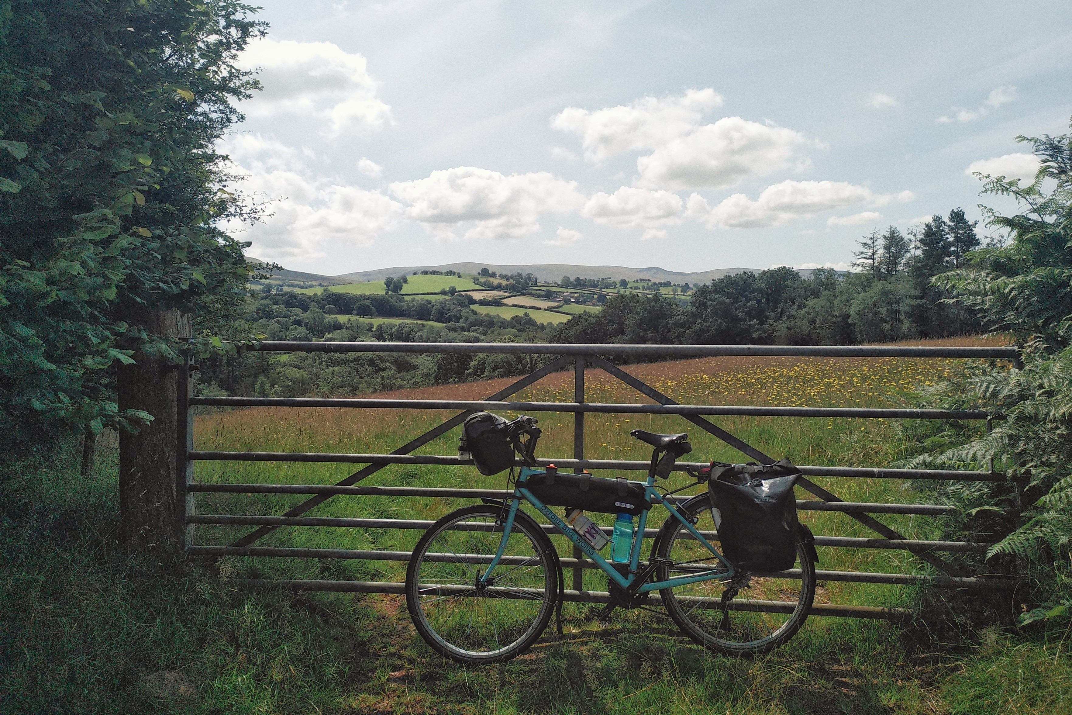 A horizontal colour photograph of a bicycle by a metal gate. In the distance there are green hills.