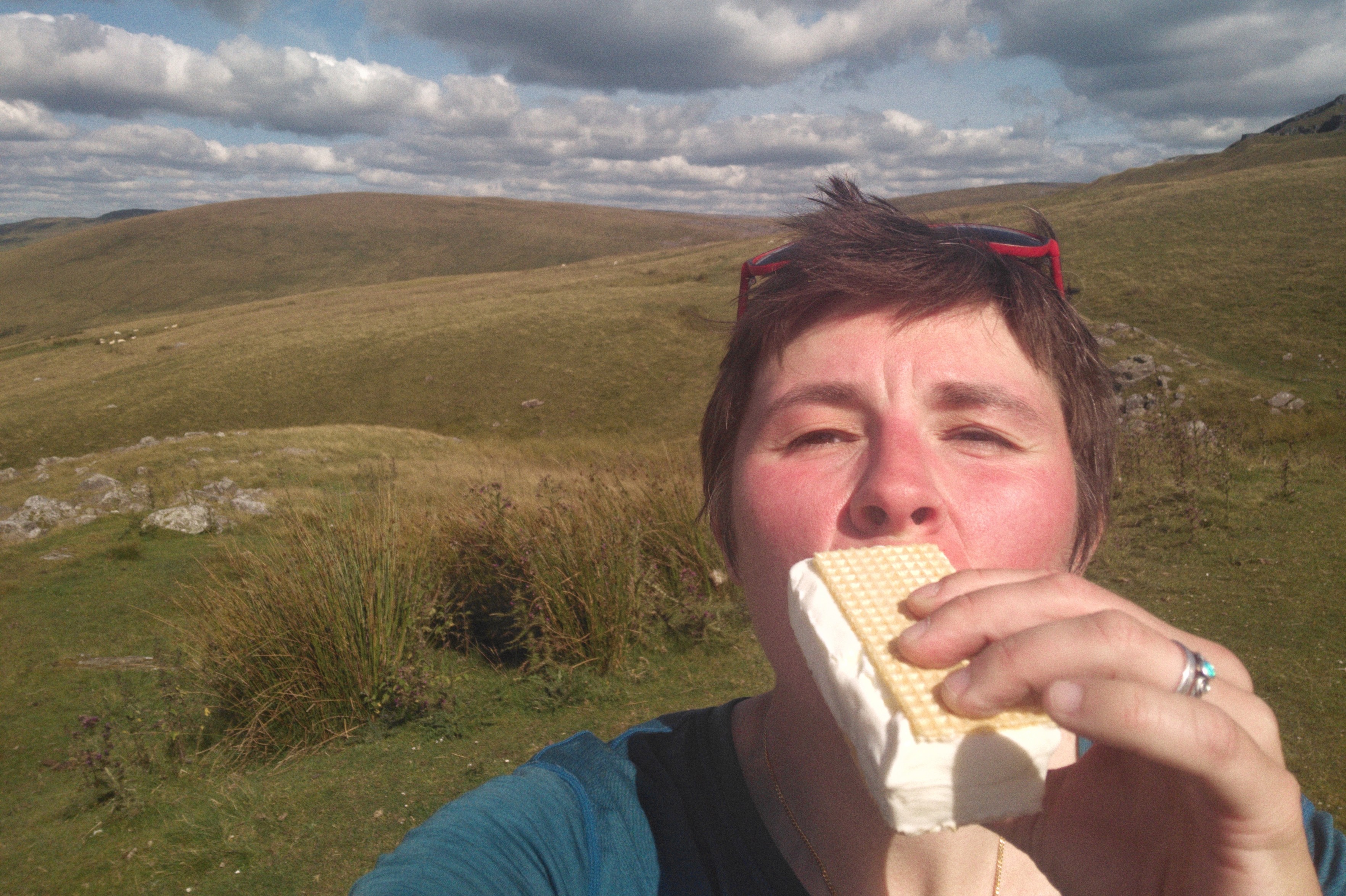 A horizontal colour photograph of me (a white person with short dark hair) with an ice-cream by my mouth. Behind me are green hills or Y Mynydd Du.