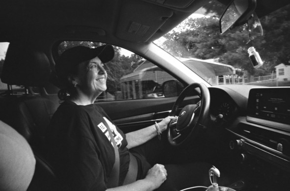 Black and white photo of a woman (my wife) wearing a baseball cap, driving her car. 

The photo is taken from the passenger seat with a wide angle lens. She's grinning for the camera, while keeping her eyes on the road as we head out for a weekend photo adventure.