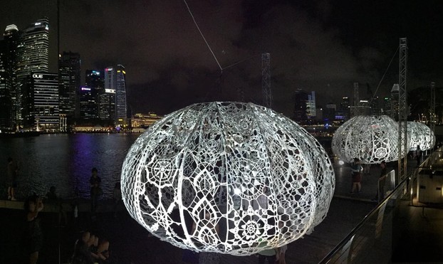 three large crocheted bulbs dangling above water in front of a city skyline