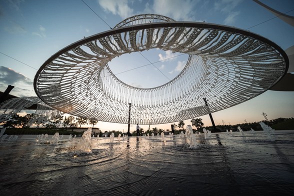 a large round hoop sculpture hangs over a small fountain. white crochet covers it