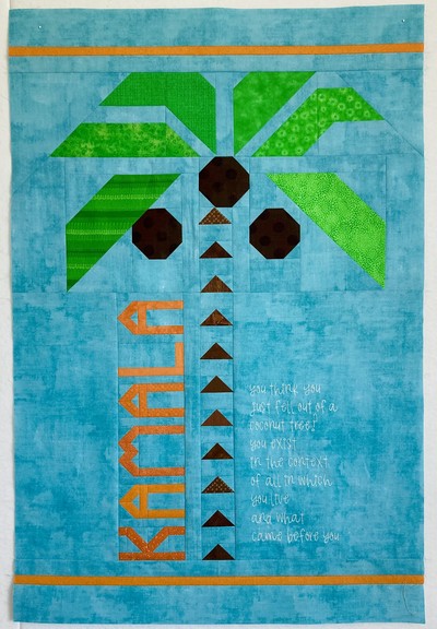Pieced Kamala Harris coconut quote quilt top shown in full. A coconut tree with scrappy green fronds on a sky blue background is the main design; the trunk of the tree is made up of equally spaced HSTs in medium brown. There are three dark brown coconuts. To the left of the trunk are scrappy orange letters with pointy tops like the HSTs that spell Kamala. To the right of the truck, her famous quote is bean-stitch embroidered in white. It reads “You think you just fell out of a coconut tree? You exist in the context of all in which you live and what came before you”