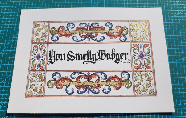 'You Smelly Badger' calligraphed in black ink blackletter script, surrounded by a decorative border of leaves and floral patterns painted in red, green, and blue, with gold and bronze metallic bits.