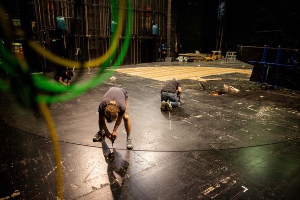 Zwei Personen arbeiten an den Bodenplatten der Bühne, hinten sieht man die offene Unterkonstruktion der Bühne aus Holz