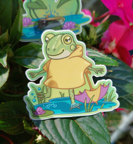 Photograph is a sticker resting on a plant. The sticker depicts a frog in a yellow raincoat splashing in a puddle with an umbrella, lilypads and some grass around the puddle