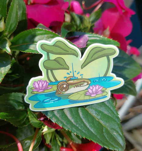 Photo of a sticker resting on flowers. The sticker depicts a frog poking its head above water with a raindrop hitting its nose. It is surrounded by water lilies and plants in the water.