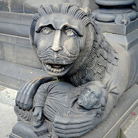 A lion (probably) sculpture with a large flowing mustache and a very human-like brown holding a human who looks drunk (he is holding a wine cup in his hands) in his unusually large hand/pawns.
