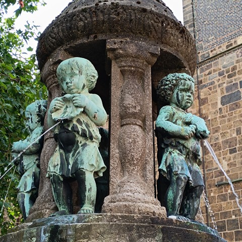 Bronze fountain with three little children in tunics, one is holding a startled frog, forever in that position, puking a stream of water.