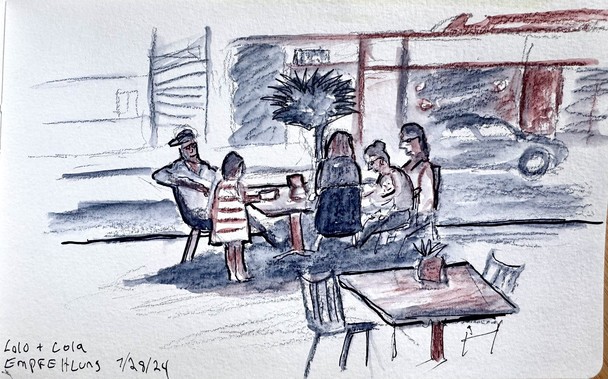 A family of six sitting at a table by the street. Everyone is seated except a young girl in a striped dress who is standing while eating. In the background vaguely shapes of city buildings and cars are visible.