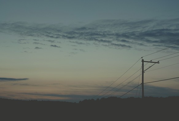 Evening sky photograph of a sunset, going from blue at the top to pale orange at the horizon, with grey horizontal clouds in the middle and close to the horizon. An electric line is seen across the sky on the right side, with the dark silhouette of an electric pole.