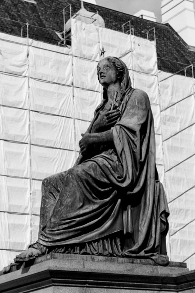 The black and white image shows a statue of a seated woman draped in robes, holding a cross to her chest. She appears to be looking upwards, possibly in a contemplative or prayerful pose. The background includes a building covered in scaffolding and protective sheeting, indicating construction or renovation work.