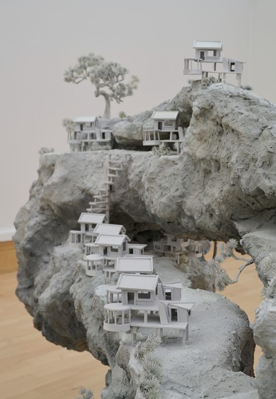 a detail of a gray sculpture with rough forms topped with small houses and trees