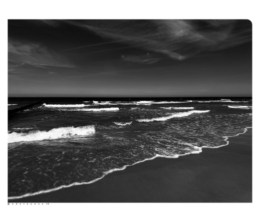 Schwarzweißfotografie: weitwinkelige Aufnahme von einem breiten Strandabschnitt, der Himmel ist grau verwaschen, die in verschiedenen Abständen anlandenden Wellen sind schaumig weiß, die Ostsee dunkel, sowie auch der dunkelgraue Sandstrand im Vordergrund 