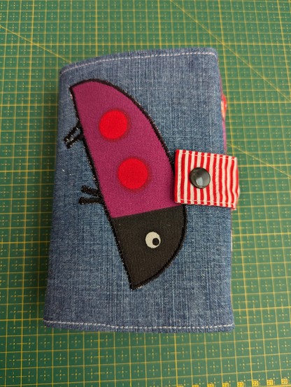 Outside of a folded travel sewing case. It's made of a blue denim fabric, has a red and white striped closing loop (with a black colour snap as a fastening) and on the blue denim a picture of a purple ladybug has been appliqued. It's about 7 inches tall and 5 inches wide when folded.
