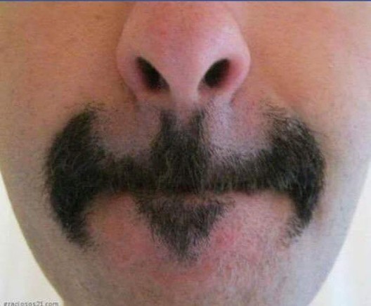 A white person with dark facial hair with their lips pressed together. The hair is shaved into an elaborate mustache with a soul patch resembling The Bat Signal from Bat Man - it's a simplified outline of a bat in flight