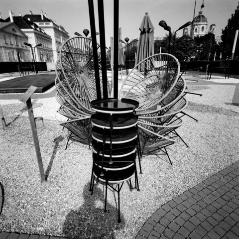 The image shows a stack of circular, woven chairs and tables arranged in a public outdoor space. The chairs are neatly piled together, creating an intricate pattern with their circular backs. In the background, there are closed umbrellas and surrounding buildings, one of which appears to be a historical structure with a dome. The scene is captured in black and white, adding a sense of contrast to the urban setting. 