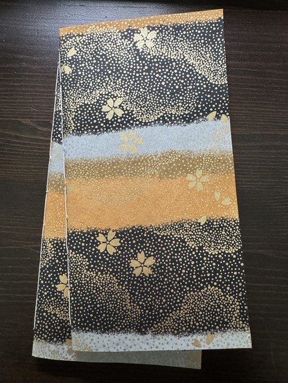 Two notebooks are stacked on a desk. Their covers are printed with orange, blue, and black horizontal bands, with a layer of speckling and small cherry blossoms in gold over top.