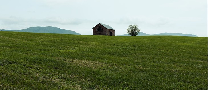 An old red barn, one wall partially decayed, stands by a tree atop a sweeping, green field. Low-lying mountains rise in the distance.