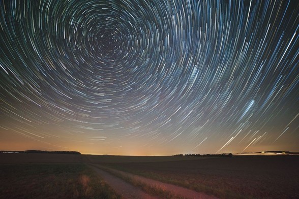 Long exposure photo of stars leaving trails while rotating around the celestial pole like a carneval ride. A soft glow from the horizon mirroring the light of some distant villages. 