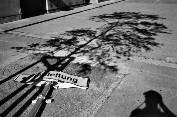 The image shows a fallen street sign lying on a sidewalk. The sign reads “Umleitung” (German for “detour”). A tree’s shadow is cast onto the pavement, and a person’s silhouette is visible in the bottom right corner. The scene is in black and white, adding a stark contrast between light and shadow.