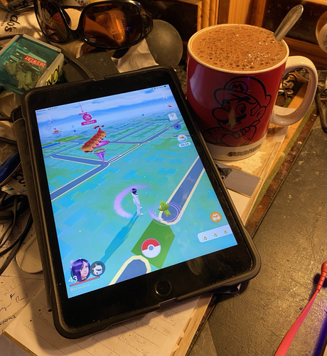 An iPad with Pokemon GO open, next to a red Mario mug full of hot chocolate.