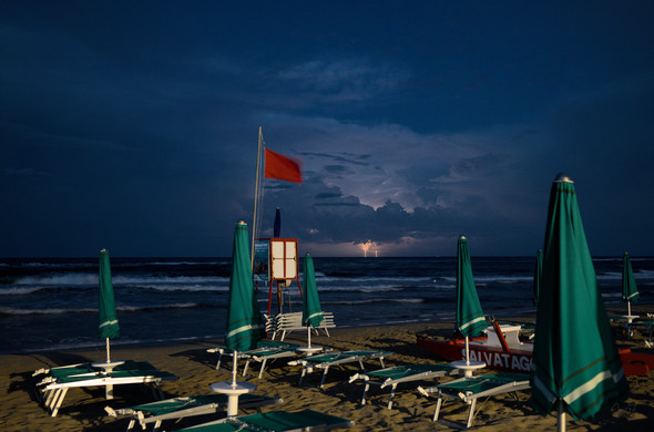 Photo of the beach in the early evening.
There are green umbrellas and a red flag waving in the wind.
Over the sea a storm is raging and a lightning strikes the sea.