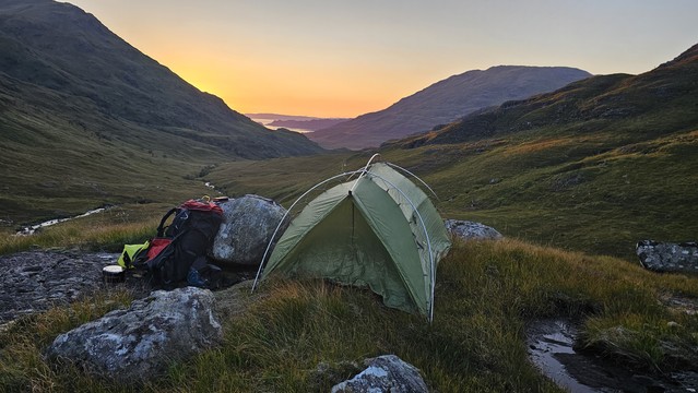 Green camping tent set up in a scenic mountain landscape during sunset, with backpack and outdoor gear on rocky terrain