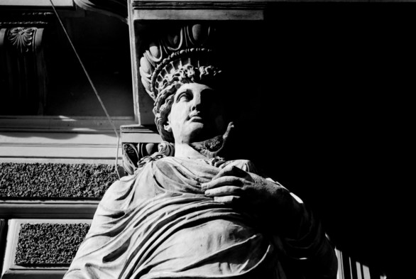 This black and white image features a close-up of a classical stone statue of a woman. The figure wears a draped garment and a decorative headdress, gazing forward with a calm, stoic expression. Her hand rests on her chest with a dove sitting on it, and the statue is partially illuminated, casting shadows that highlight its intricate details.