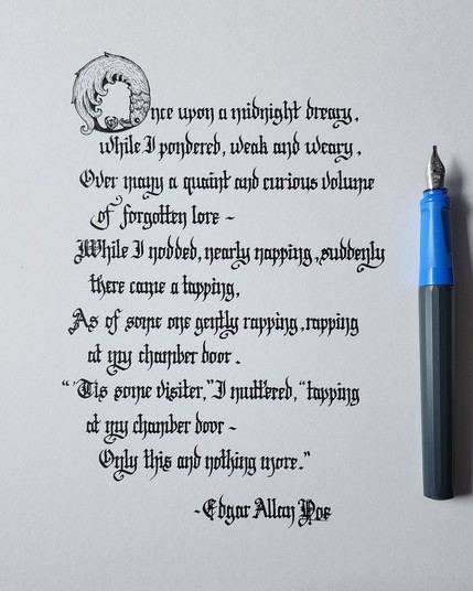 A verse from Poe's poem 'The Raven', calligraphed in black ink blackletter script, with an ornate first 'O' that's illustrated like a raven curling in on itself;

Once upon a midnight dreary, while I pondered, weak and weary,
Over many a quaint and curious volume of forgotten lore -
While I nodded, nearly napping, suddenly there came a tapping,
As of some one gently rapping, rapping at my chamber door.
