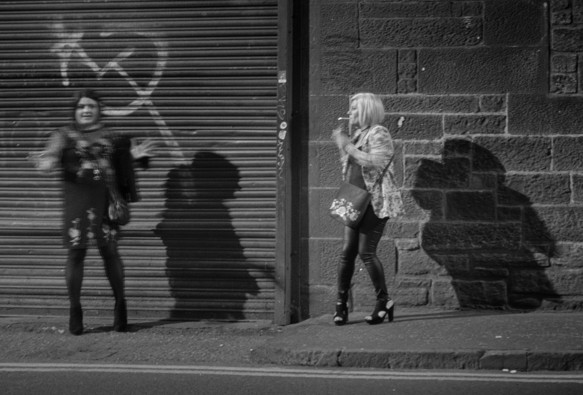 A black and white photo taken at night in The Cowgate part of the Old Town. Two women are on a night out, caught in the bright streetlights they cast strong shadows on the wall and shuttered entrance behind them. At left, one woman is moving and gesticulating, slightly blurred by motion, at right, her companion it turned towards her with the bright end of her lit cigarette a light smudge. Part of the Edinburgh Nights series. Edinburgh, Scotland.

Photo by and copyright of Lynn Henni.